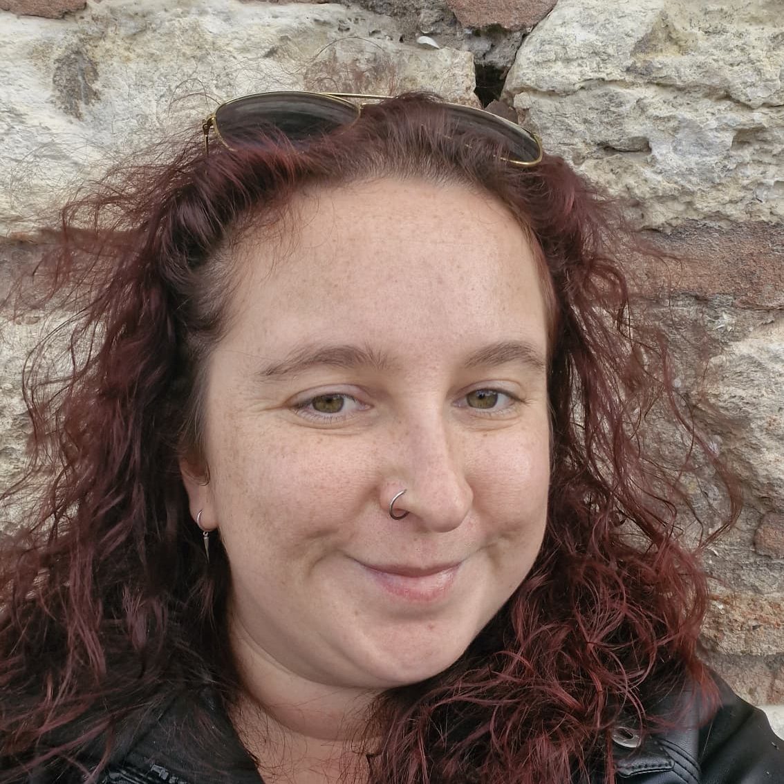 Heather has red curly hair, and a nose ring and is smiling at the camera